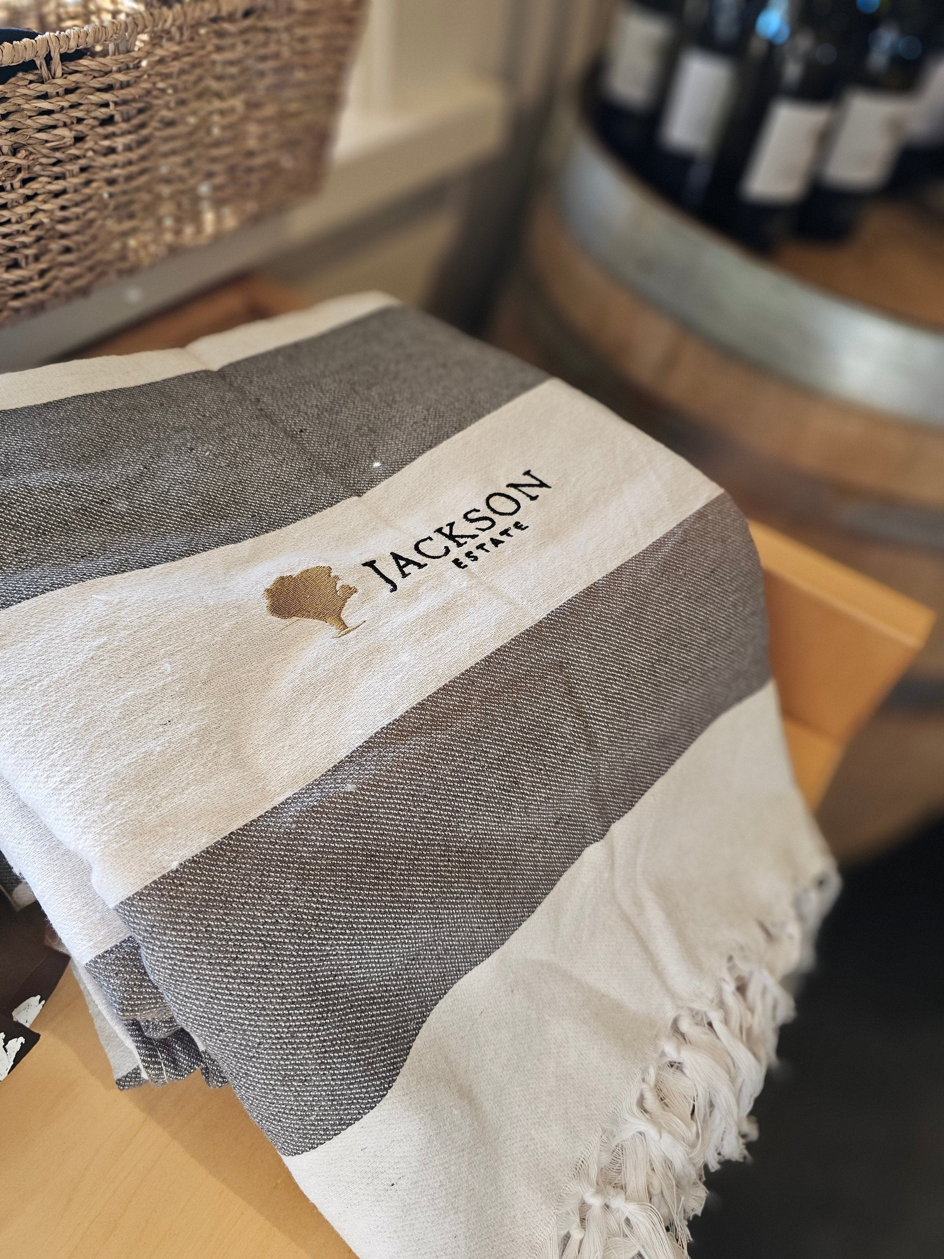 Jackson Estate branded striped cotton throw with fringed edge, displayed on a wooden surface with wine bottles in the background