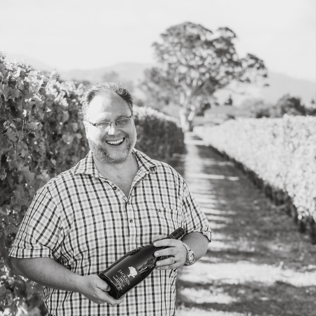 Man smiling and holding a wine bottle while standing between vineyard rows, hills visible in the background