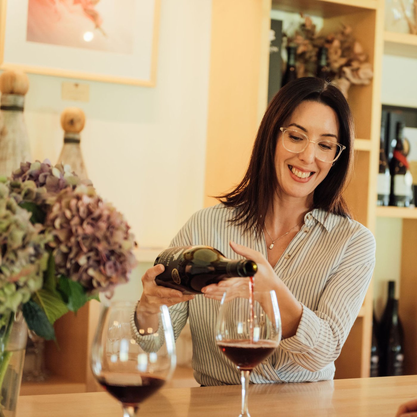 Smiling cellar door host pouring red wine at a tasting counter with hydrangeas and wine glasses nearby