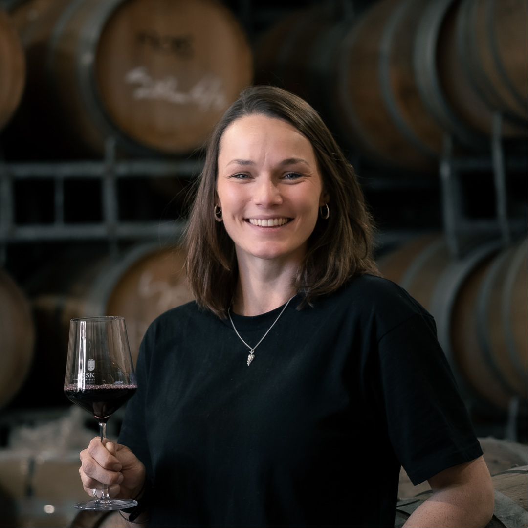 Woman holding a glass of red wine and smiling, standing in front of oak barrels in a winery barrel room.