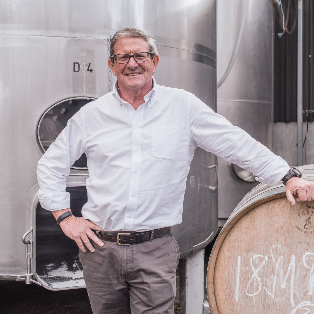 Man leaning on an oak wine barrel in front of stainless steel fermentation tanks at a winery
