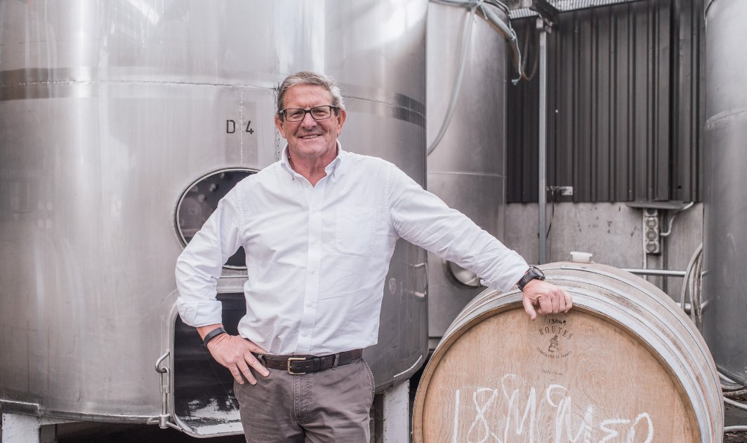 Man leaning on an oak barrel in a winery, with large stainless steel fermentation tanks behind him