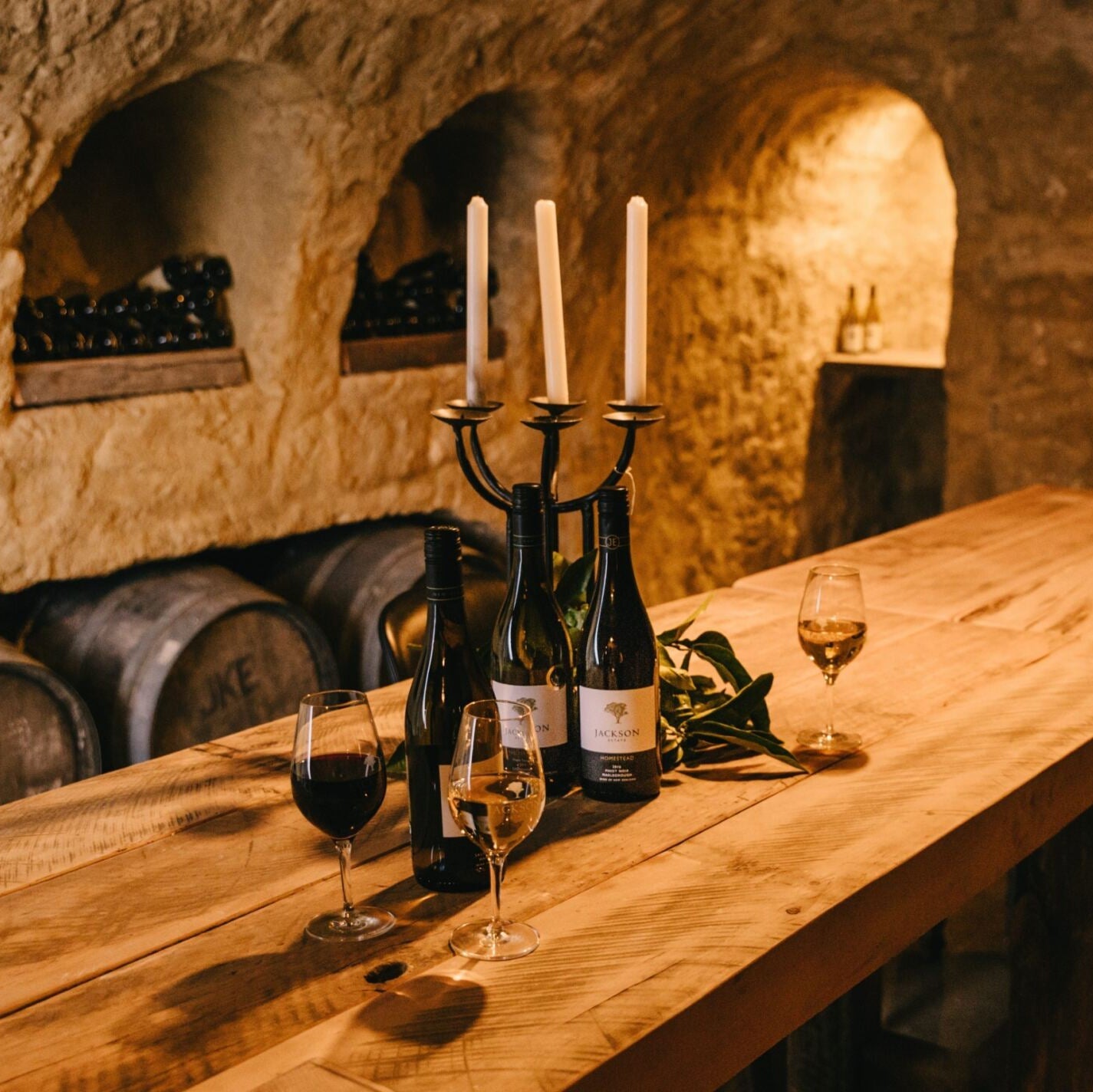 Three Jackson Estate wine bottles and filled glasses on a wooden table in a candlelit stone wine cellar with oak barrels