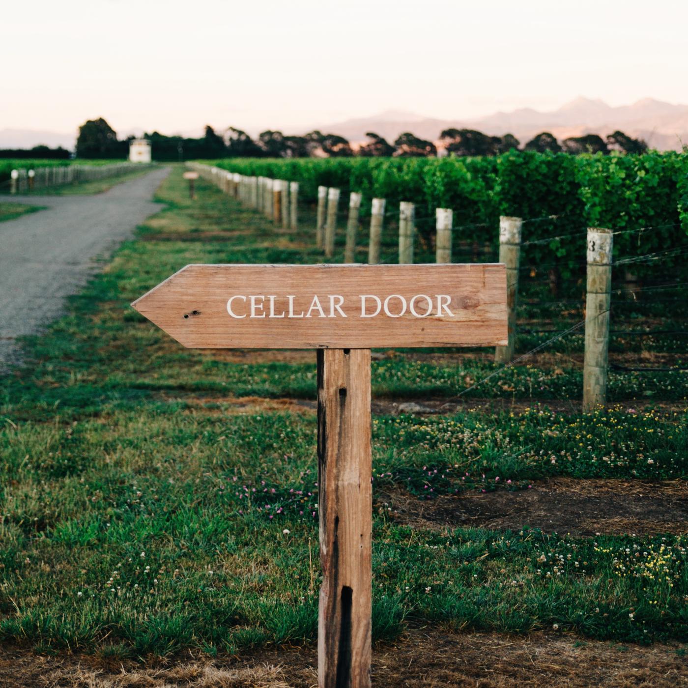 Wooden "Cellar Door" directional sign beside a vineyard lane at dusk, with rows of vines and mountains in the background.