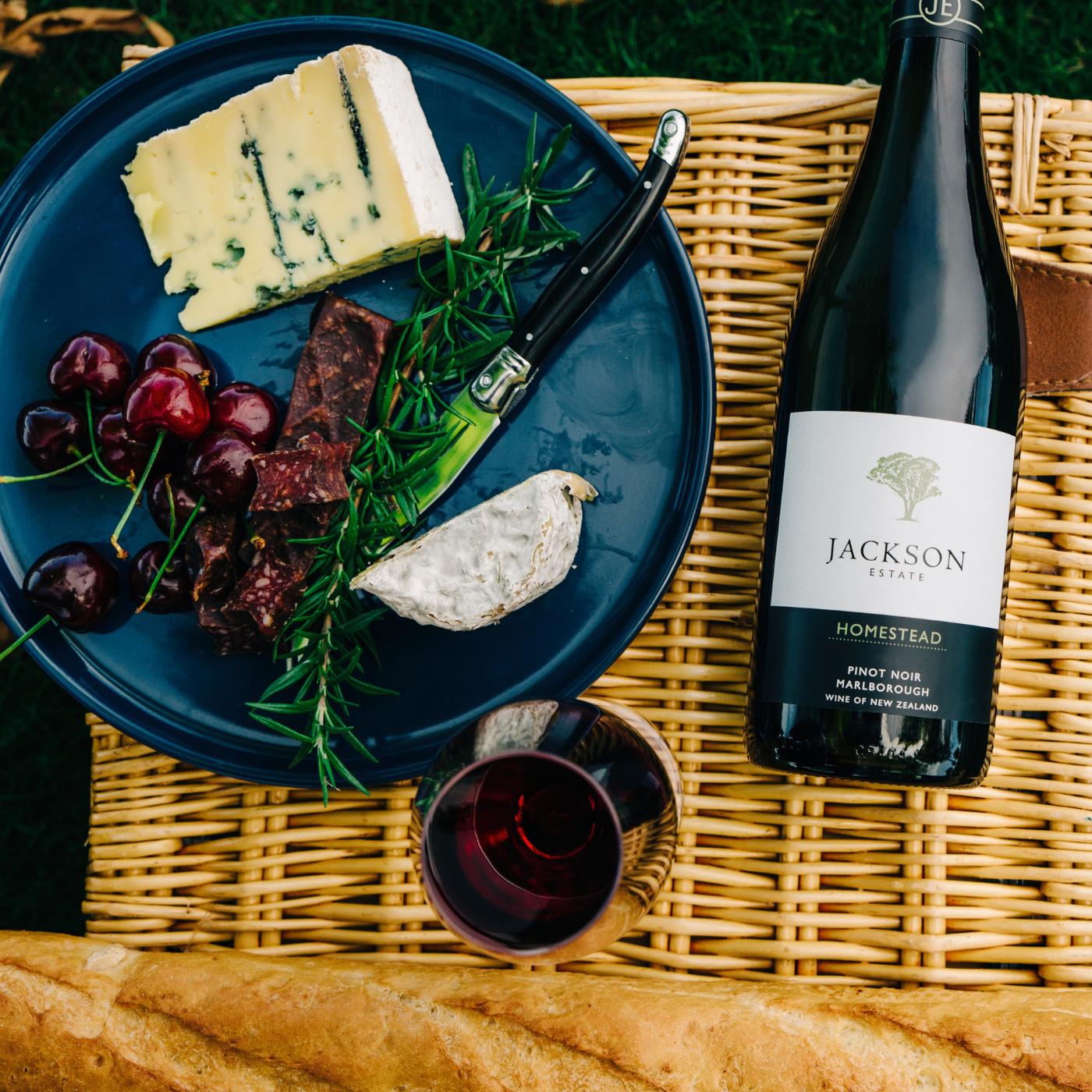 Jackson Estate Homestead Pinot Noir bottle beside a cheese and charcuterie plate with cherries and a glass of red wine on a picnic basket