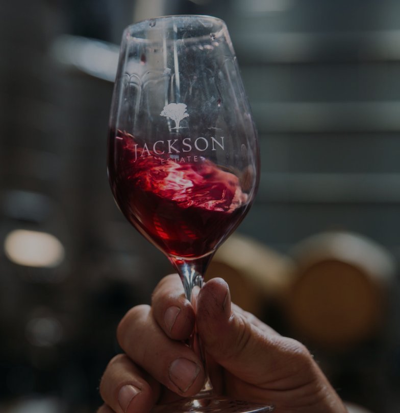 Hand holding a Jackson Estate branded wine glass with swirling red wine, oak barrels softly blurred in the background