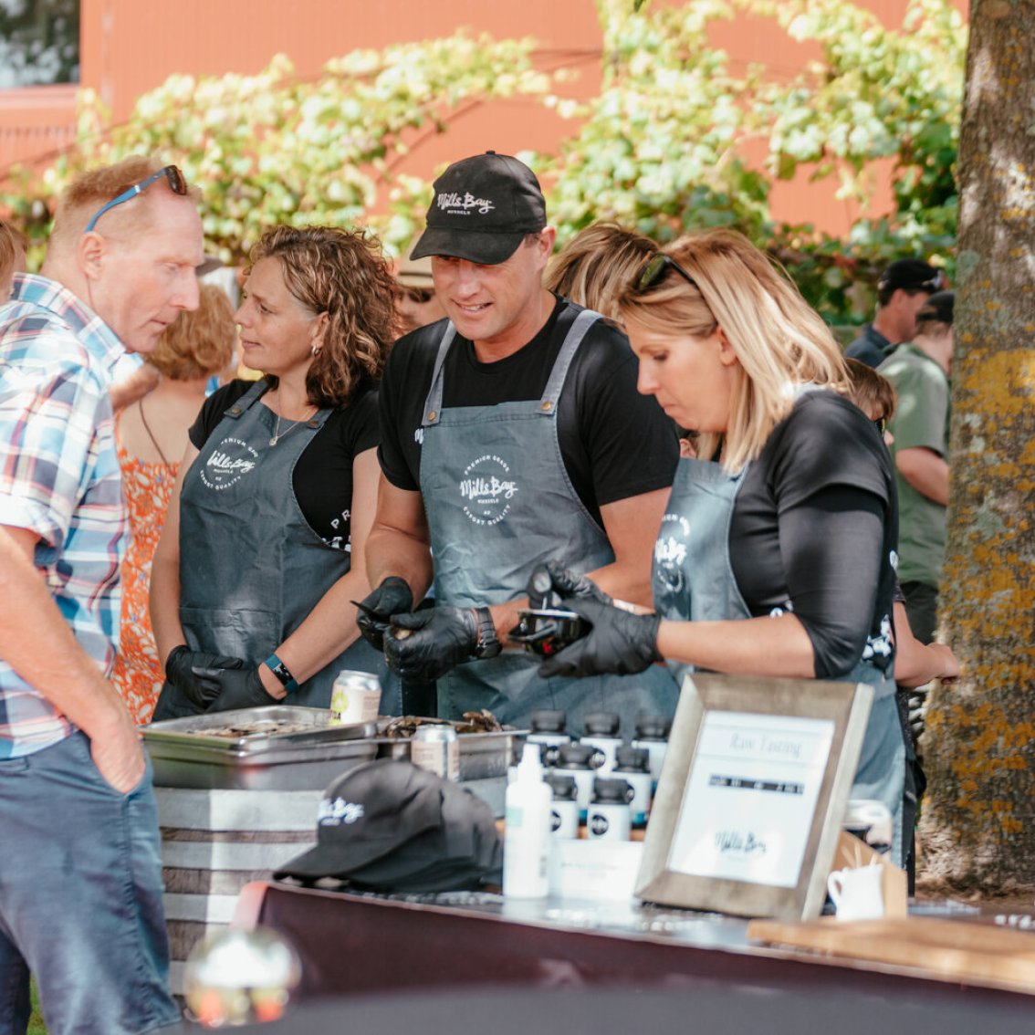 Staff in branded aprons serving guests at an outdoor food and wine event on a sunny day