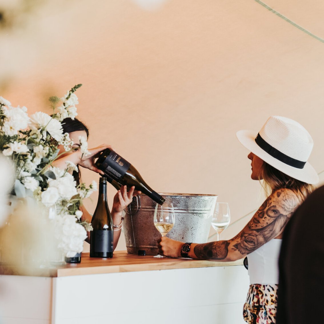 A staff member pours wine for a guest at an outdoor tasting event, with white flowers, wine bottles, and an ice bucket on the counter.