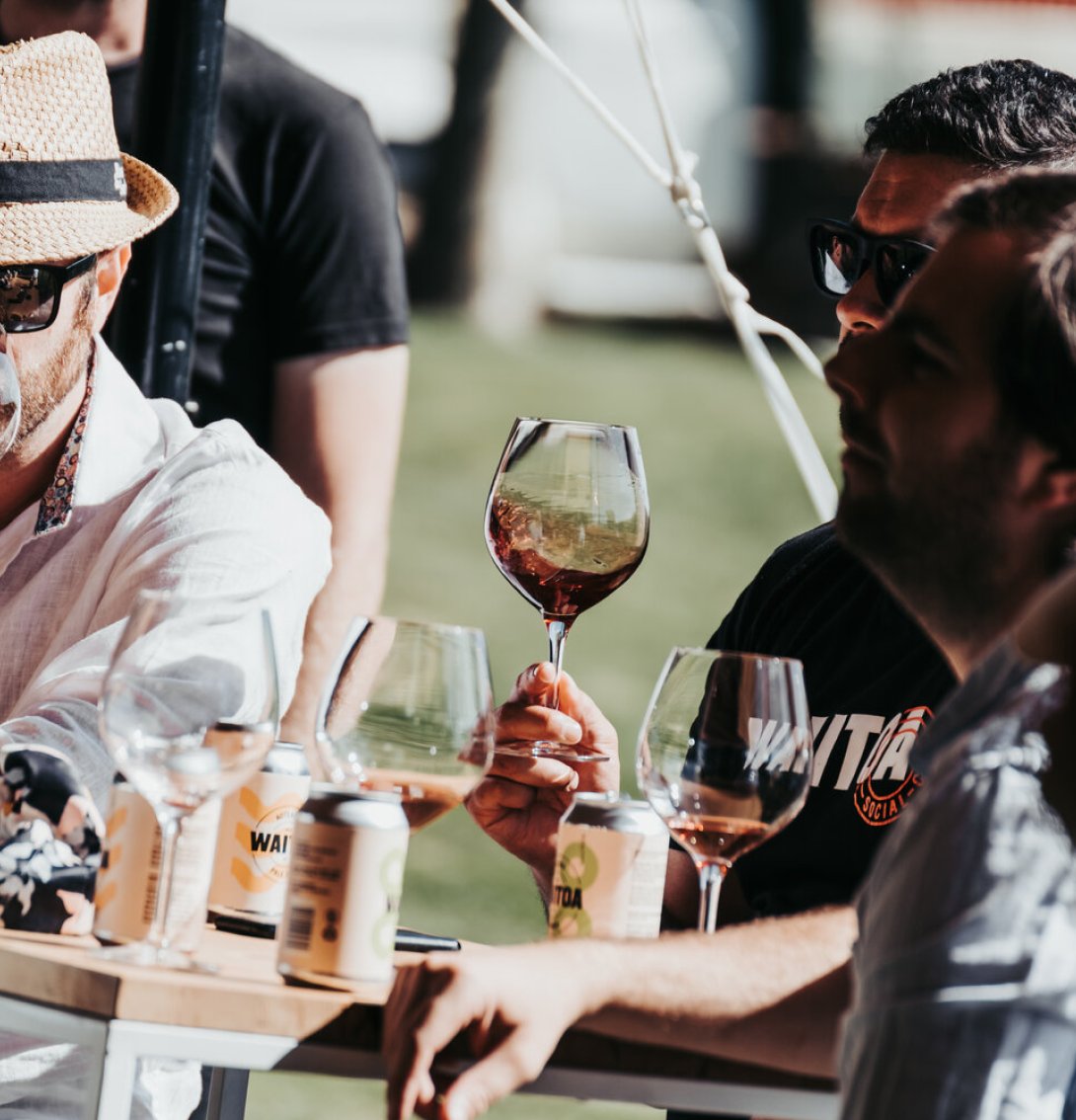 Two men examining wine glasses at an outdoor tasting event, with cans and several wine glasses on the table beside them