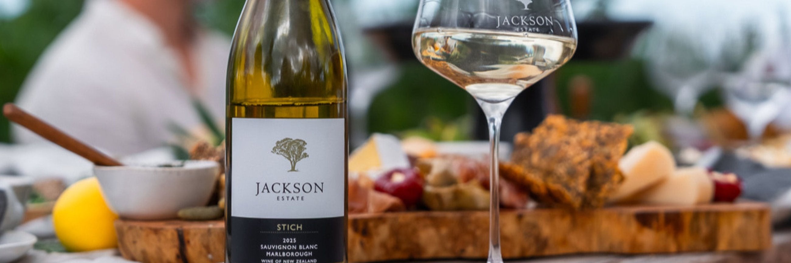 Jackson Estate Stich Sauvignon Blanc 2025 beside a glass of white wine and a charcuterie and cheese board at an outdoor table